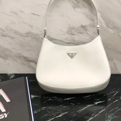 A white leather shoulder bag with a silver triangle plaque standing on a dark marble surface